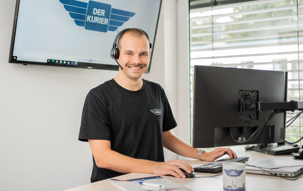Ein lächelnder Mann mit Kopfhörern sitzt am Schreibtisch und arbeitet am Computer. Im Hintergrund ist ein Bildschirm mit dem Logo "DER KURIER" zu sehen.