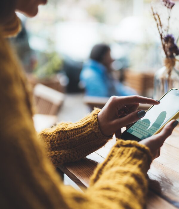 Eine Person in einem gelben Pullover nutzt ihr Smartphone, während sie an einem Tisch in einem Café sitzt.