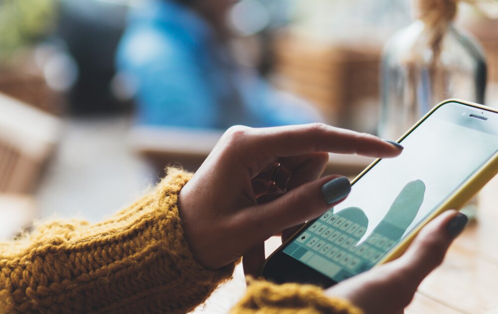 Eine Person in einem gelben Pullover nutzt ihr Smartphone, während sie an einem Tisch in einem Café sitzt.