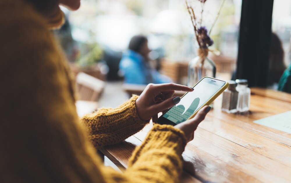 Eine Person in einem gelben Pullover nutzt ihr Smartphone, während sie an einem Tisch in einem Café sitzt.