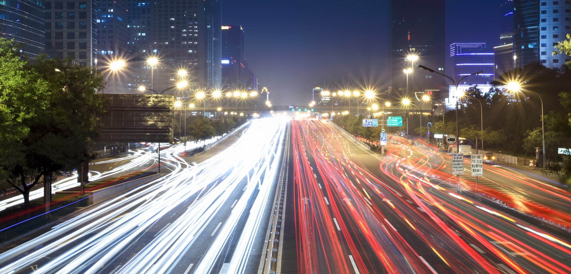 Eine stark befahrene Straße bei Nacht, beleuchtet von vielen Lichtern, mit schnell vorbeifahrenden Fahrzeugen, die rote und weiße Lichtstreifen erzeugen.