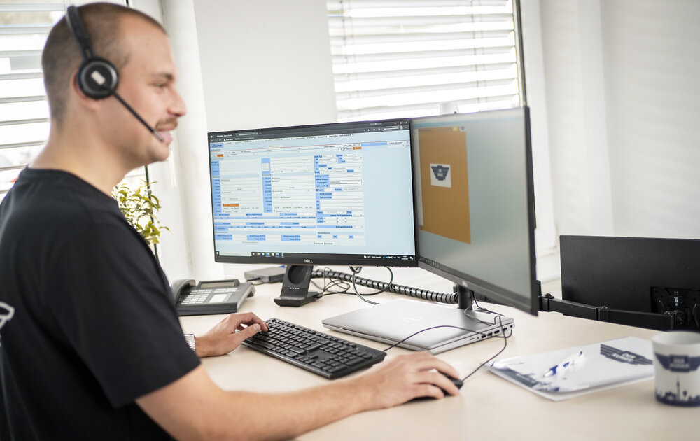 Ein Mann mit Headset sitzt an einem Schreibtisch und arbeitet an zwei Computermonitoren. Auf dem Bildschirm sind Daten und Formulare sichtbar. Der Schreibtisch enthält einen Computer, eine Tastatur und eine Tasse. Natürliches Licht strömt durch die Fenster.