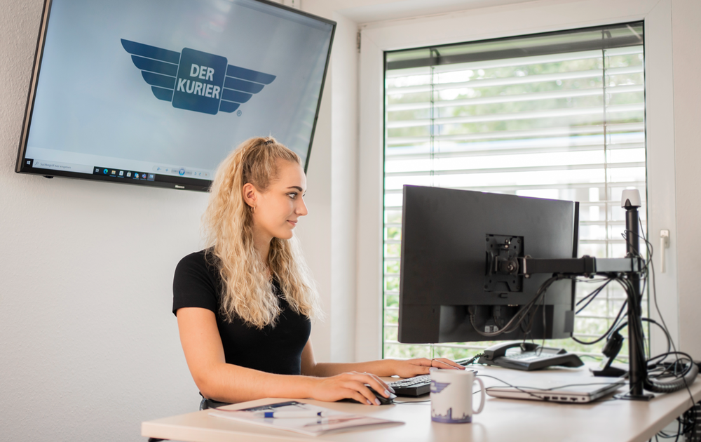 Eine Frau sitzt an einem Schreibtisch und arbeitet am Computer. Im Hintergrund ist ein Bildschirm mit dem Logo „DER KURIER“ sichtbar.