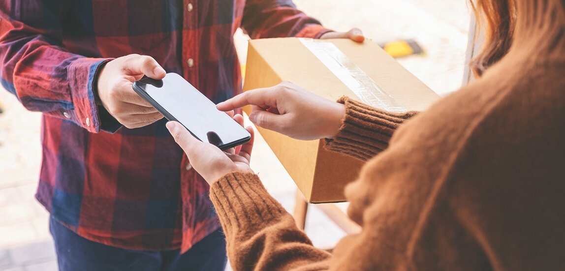 Eine Person übergibt ein Paket, während die andere ein Smartphone in der Hand hält. Es scheint sich um eine Lieferung oder Abholung zu handeln.