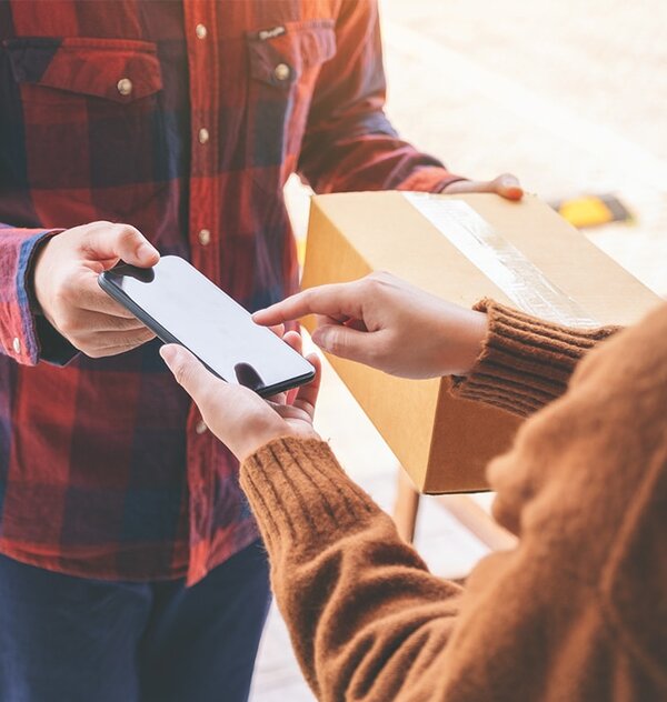 Eine Person übergibt ein Paket, während die andere ein Smartphone in der Hand hält. Es scheint sich um eine Lieferung oder Abholung zu handeln.