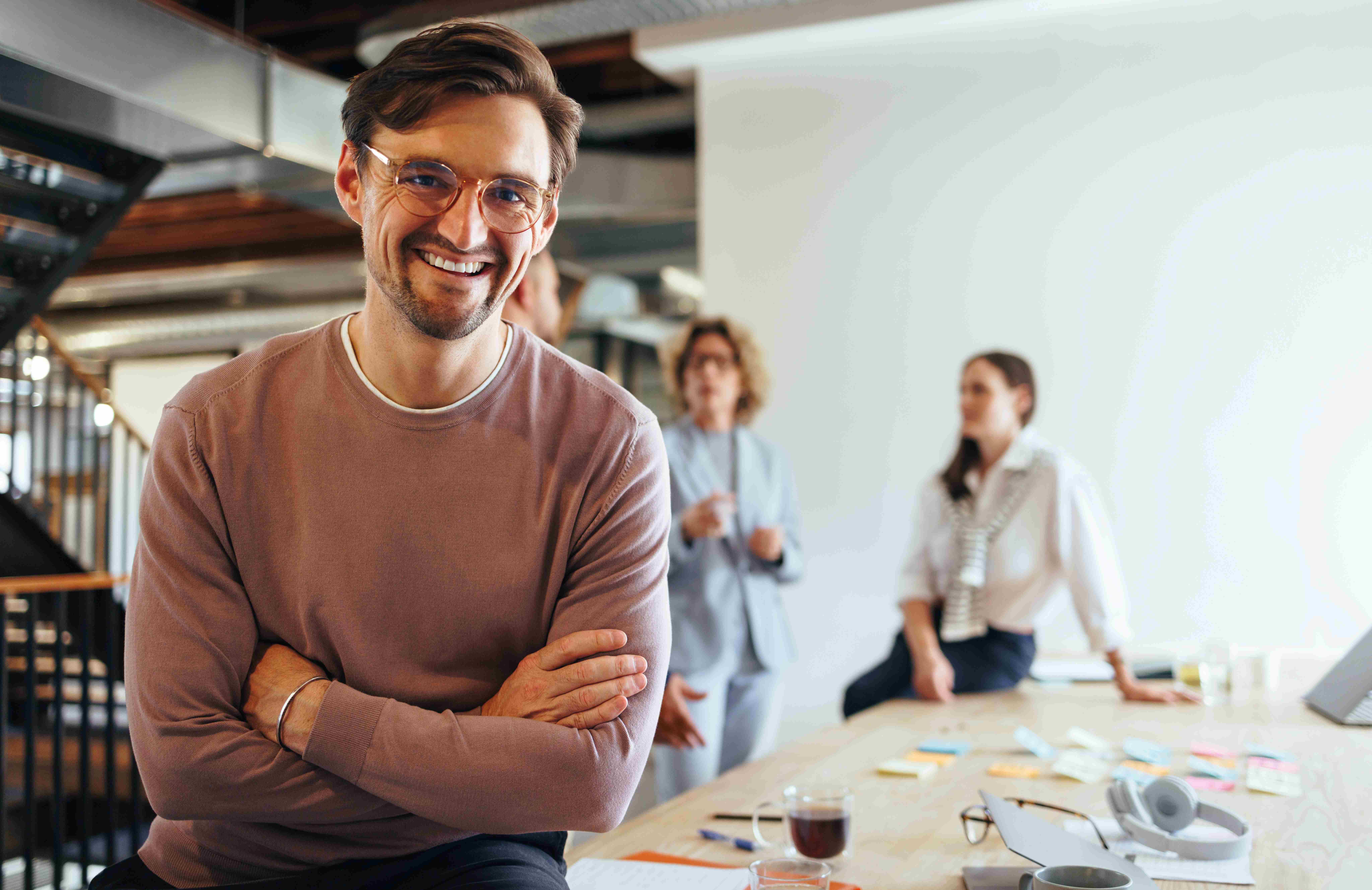 Ein lächelnder Mann mit Brille sitzt an einem Tisch in einem modernen Büro, während zwei weitere Personen im Hintergrund miteinander sprechen.